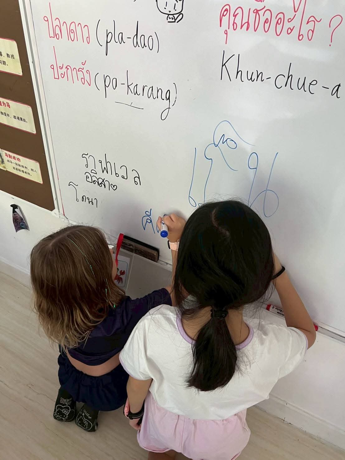 Two young children practicing writing Thai on a whiteboard